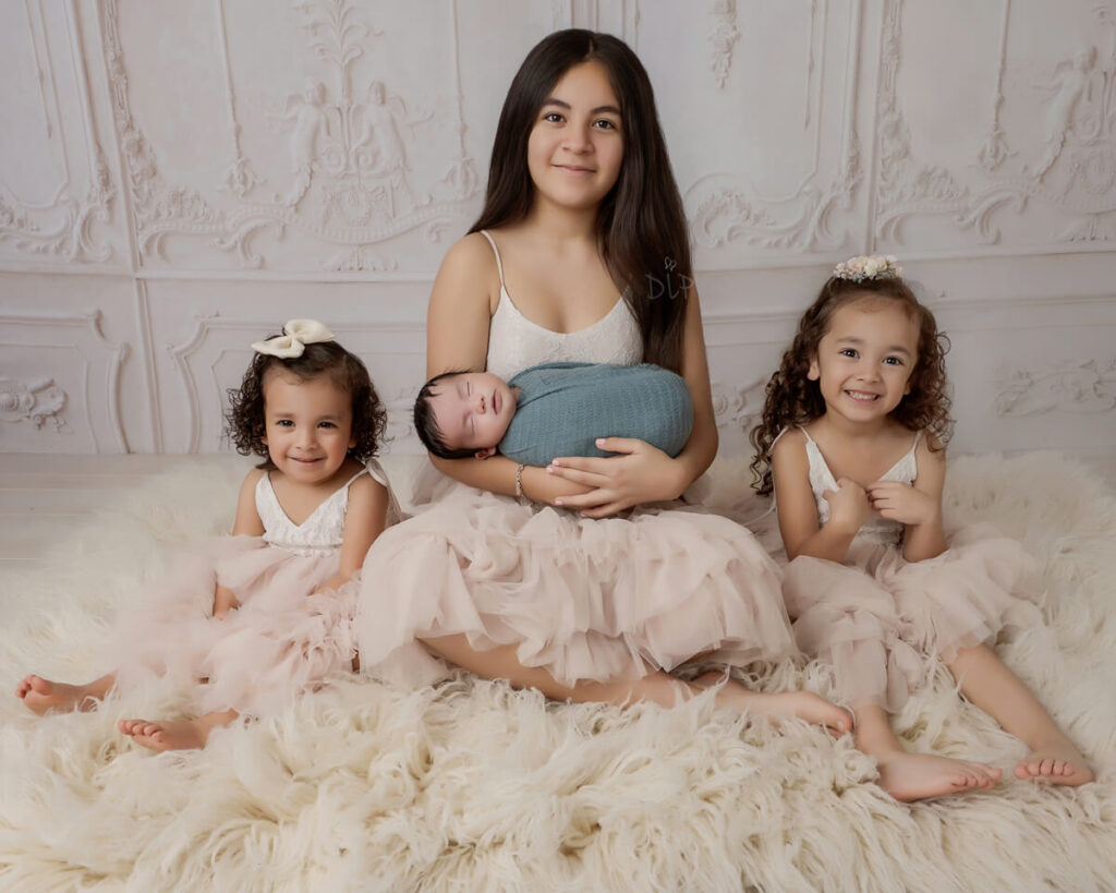 3 sisters sitting on a cream rug, the oldest is holding newborn baby brother. Image by Austin Luxury Newborn Photographer Dazzling Light Photography